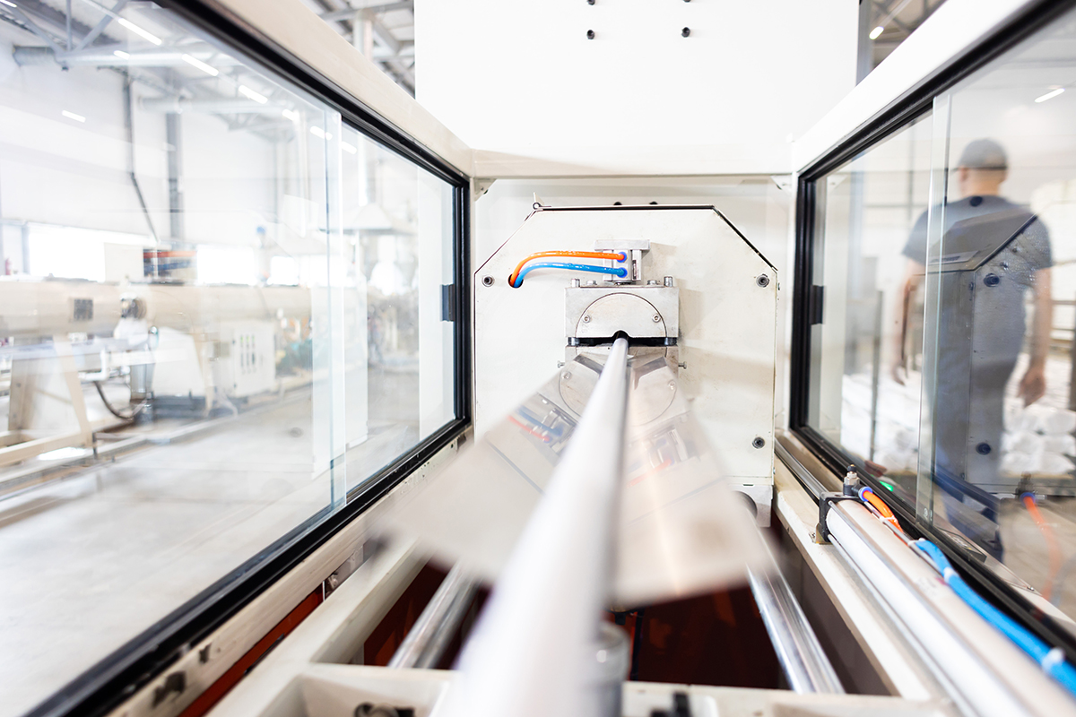 This is a view inside an extrusion machine. A plastic pipe is extruded through the inside. A worker can be seen to the right through a viewing window.