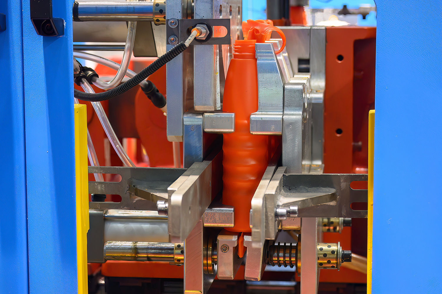 A look at a blow-molding machine creating orange bottles, with many moving parts showing signs of molding lubrication.